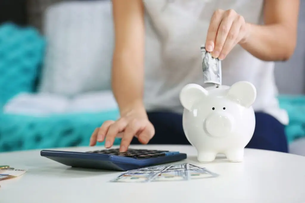 A close-up of a person placing money into a white piggy bank while using a calculator, with dollar bills spread on the table.