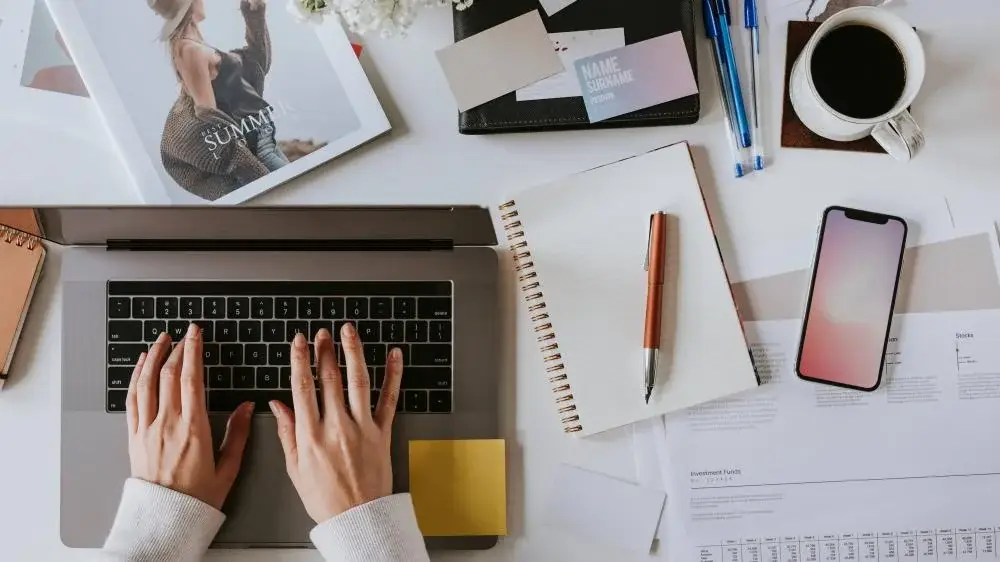 Hands typing on a laptop surrounded by notebooks, pens, a smartphone, and a cup of coffee on a workspace.