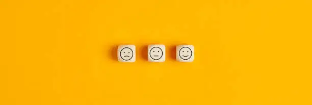 Three wooden blocks with sad, neutral, and happy faces on a yellow background representing survey responses.