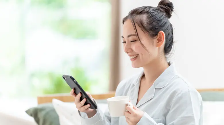 A woman smiling while using her smartphone and holding a coffee cup, demonstrating how to make money from your phone in a cozy, relaxed setting.