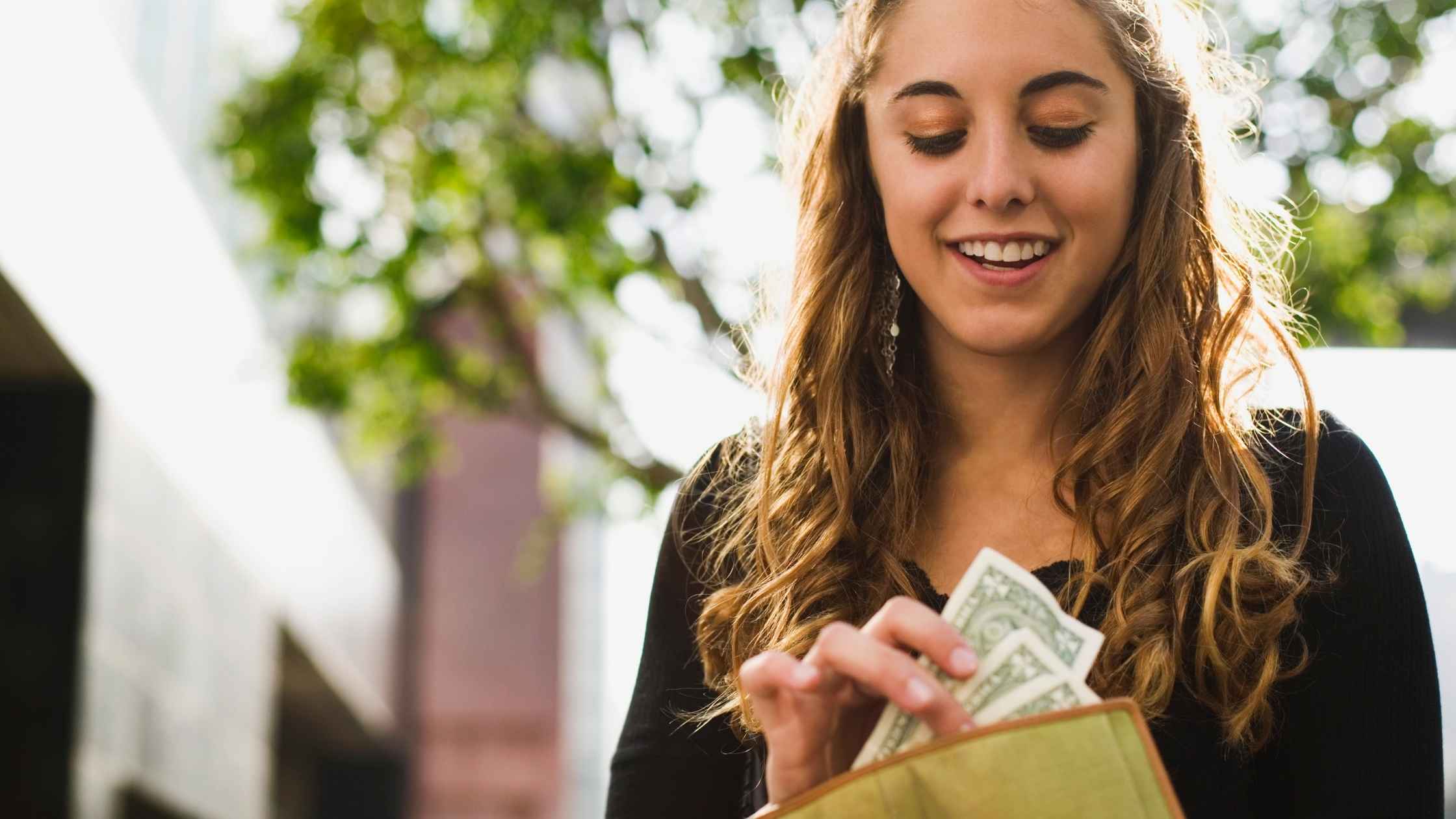 A cheerful woman holding a stack of dollar bills and a smartphone against a bright yellow background, symbolizing earning money with apps.
