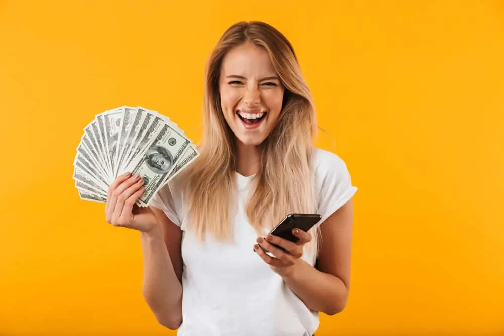 A cheerful woman holding a stack of dollar bills and a smartphone against a bright yellow background, symbolizing earning money with apps.