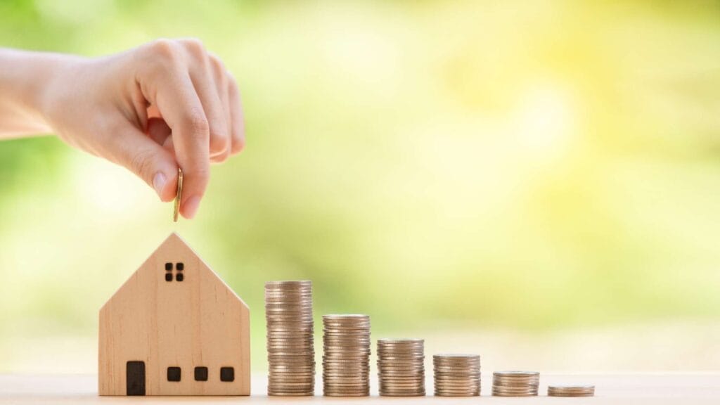 Hand placing a coin into a house model beside stacked coins, symbolizing saving for a house.