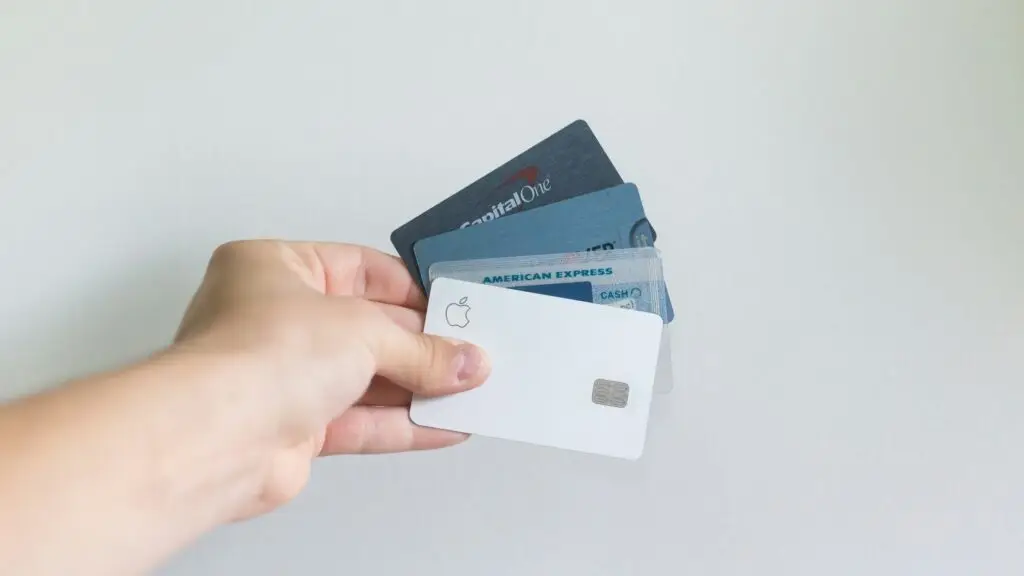 A hand holding a variety of credit cards, including Apple, American Express, and Capital One, against a plain background.