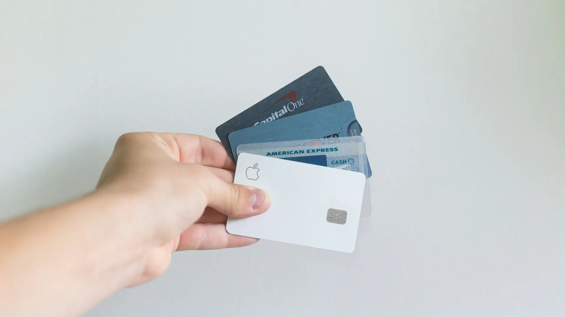 A hand holding a variety of credit cards, including Apple, American Express, and Capital One, against a plain background.