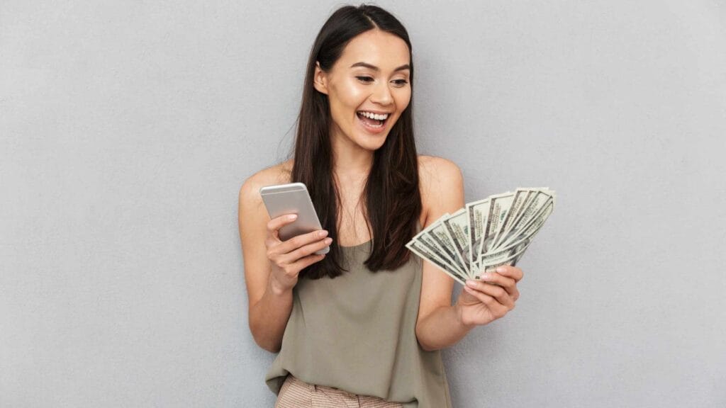 A smiling woman holding a smartphone and cash, representing ways to make money from your phone.