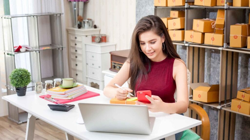 A woman working from home, packaging products with a laptop, phone, and organized supplies, showcasing a successful home business.