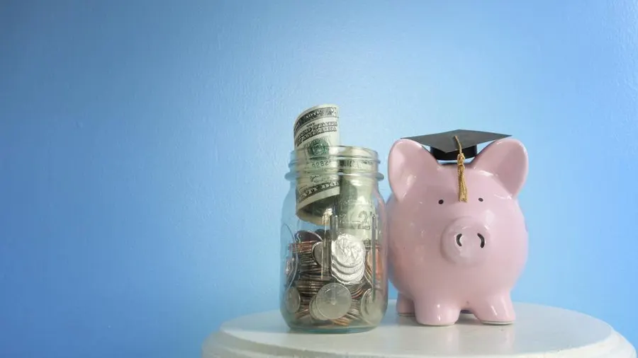 Pink piggy bank wearing a graduation cap next to a jar filled with coins and dollar bills, symbolizing saving for student loans.