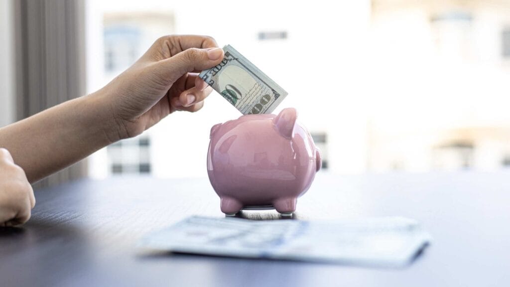 A person placing a hundred-dollar bill into a pink piggy bank on a table, symbolizing saving money.