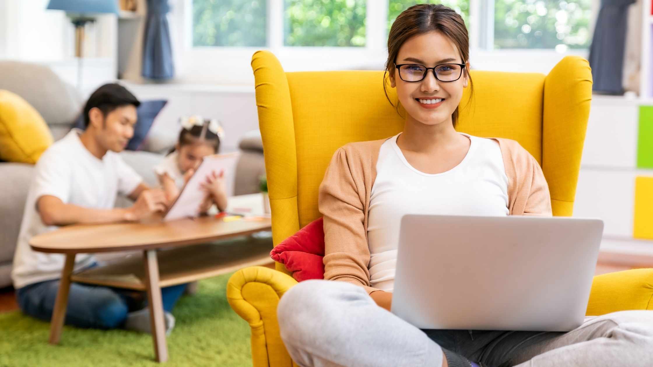 Smiling woman working on her laptop in a bright yellow chair while her family plays in the background, showcasing a work-from-home setup.