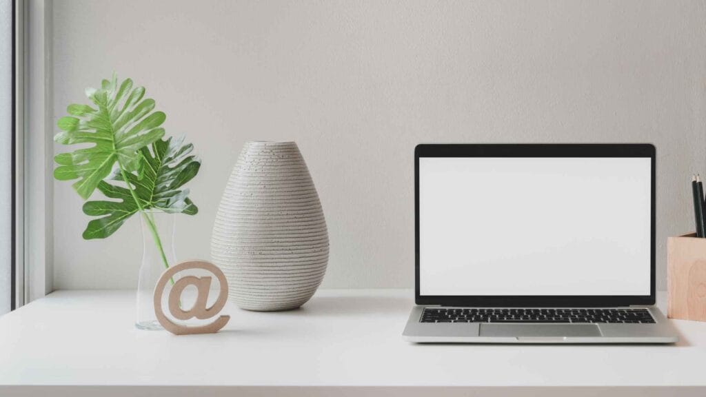 A laptop on a white desk with a decorative vase, green leaves, and an '@' symbol representing the best emailing service setup.