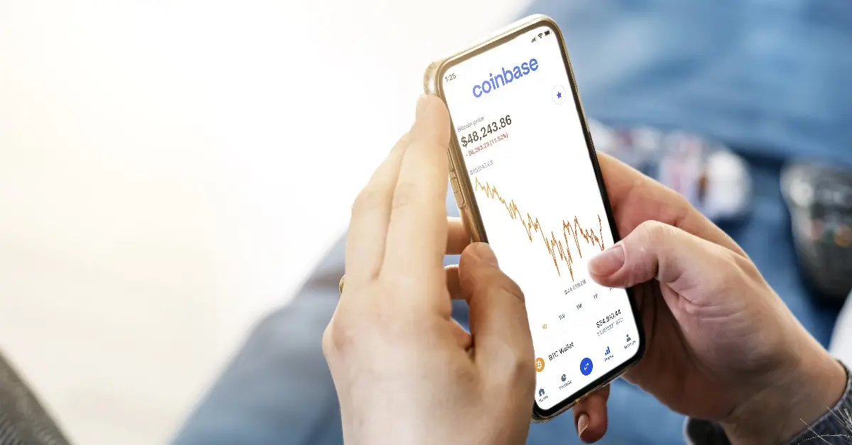 A person holding a smartphone displaying the Coinbase app with a live cryptocurrency price chart.