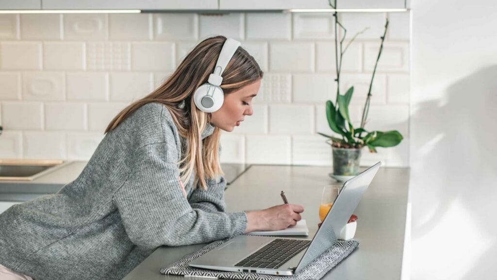 Woman wearing headphones writing notes while working on a laptop, symbolizing steps on how to create an online course.