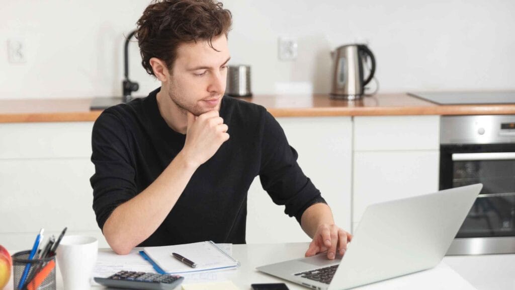 A man working on remote research jobs in a kitchen setup with a laptop, notebook, and calculator.