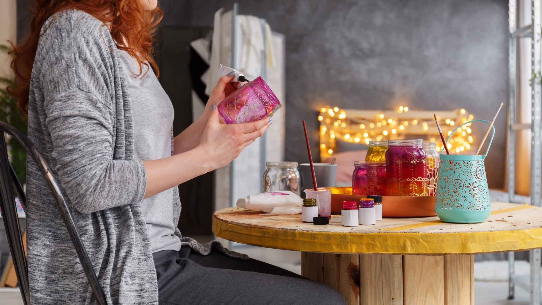 Woman painting a glass jar at a craft table, surrounded by DIY arts and crafts materials
