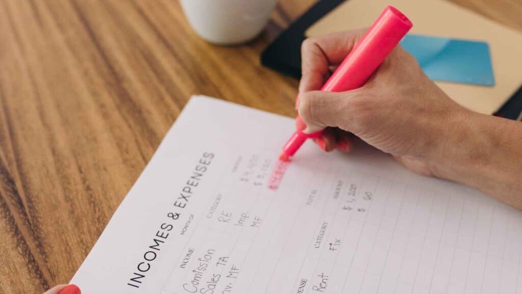 A list with a person using a sharpie to highlight their monthly income and expenses to manage finances.