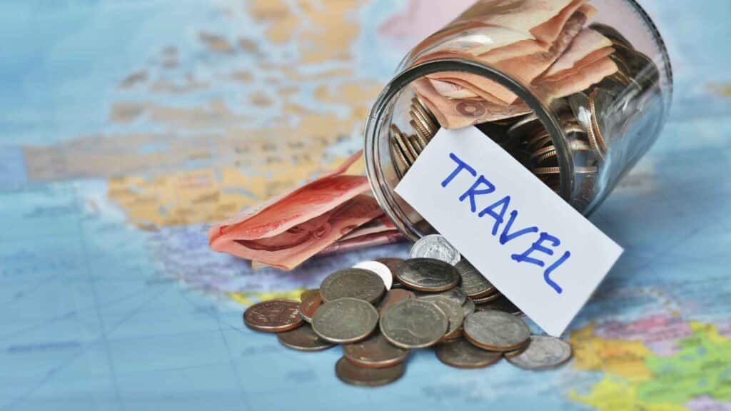 A jar spilled over with money falling out onto a map with a card that says "travel," representing planning ahead for budget travel expenses.